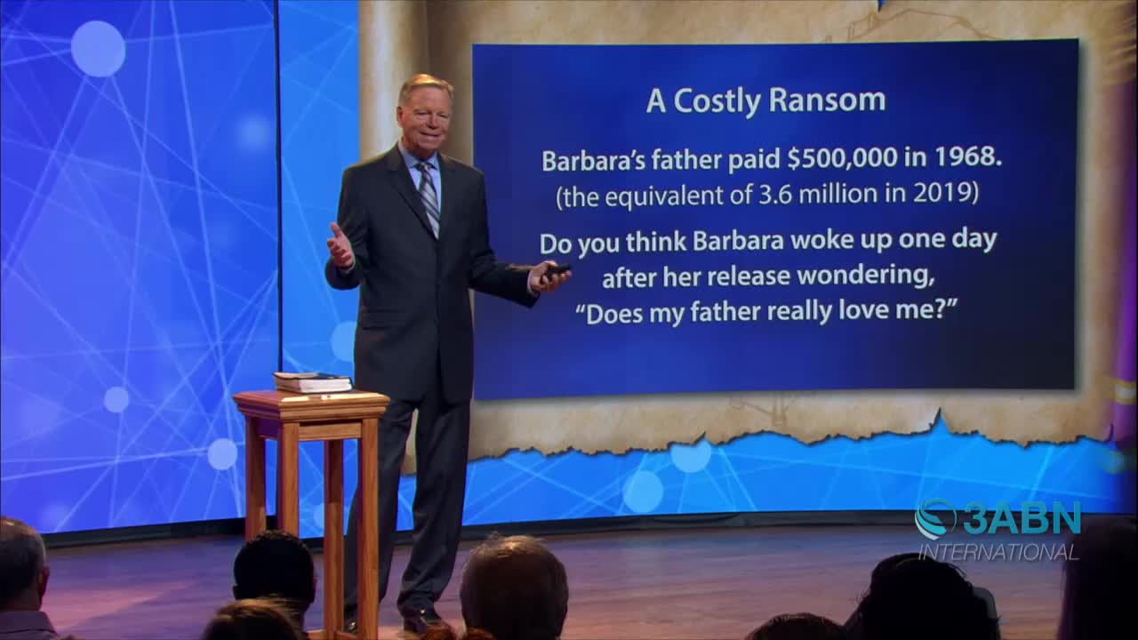On a stage for 3ABN International, a man in a suit gestures toward a screen with text about a costly ransom. He stands near a small table holding a book, addressing an audience.
