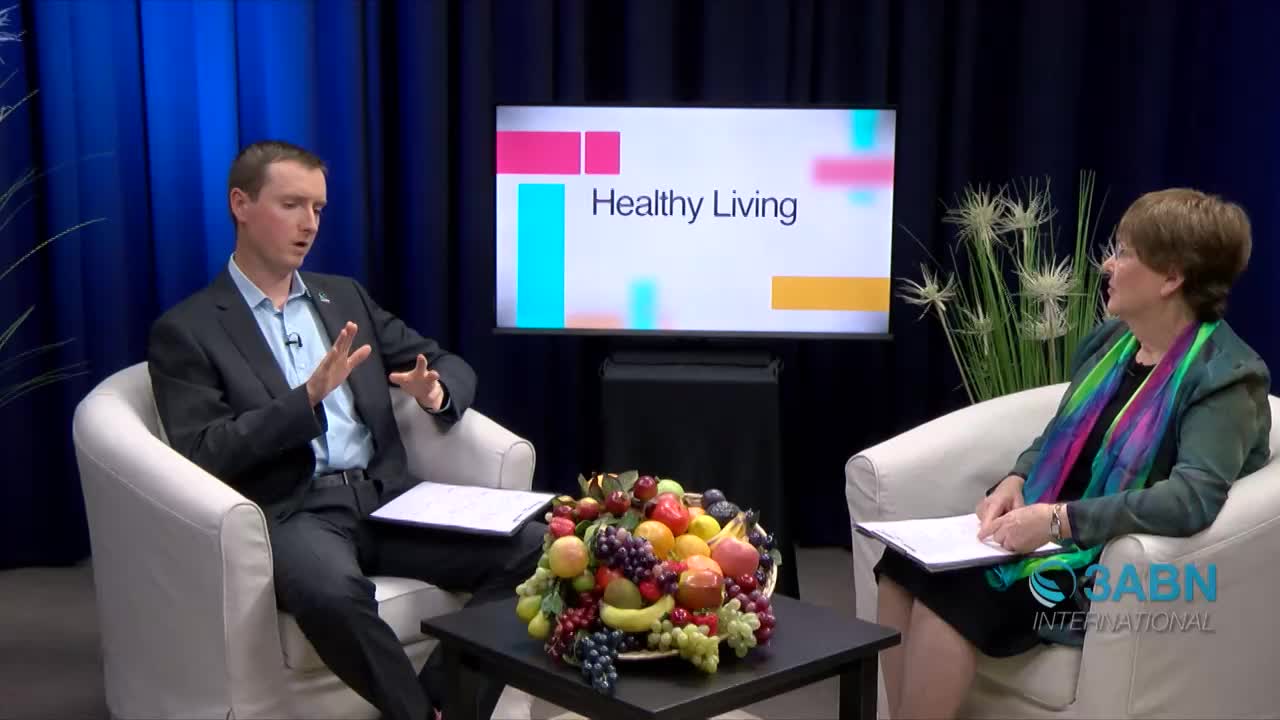 A man in a suit gestures with his hands while speaking to a woman on a "Healthy Living" segment for 3ABN International. A table between them holds a large arrangement of colorful fruit.
