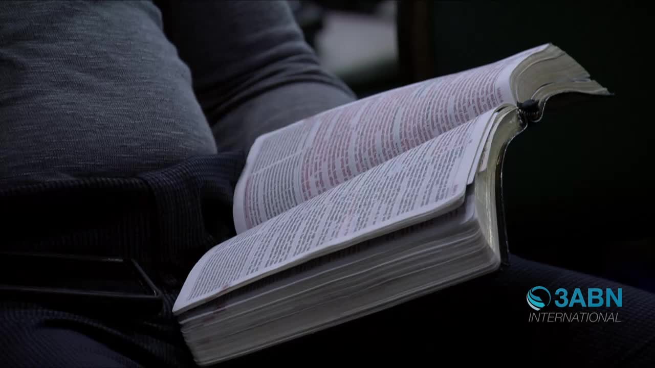 An open Bible rests in someone's lap, the pages curved and illuminated. The logo for 3ABN International is visible in the corner.
