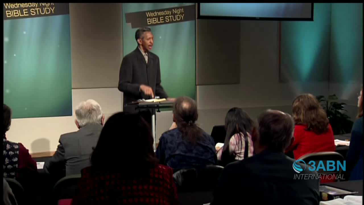 A man in a dark suit gestures while speaking at a lectern, as a group of people listen in the audience. The scene is set for a Wednesday Night Bible Study, broadcast by 3ABN International from the United States.
