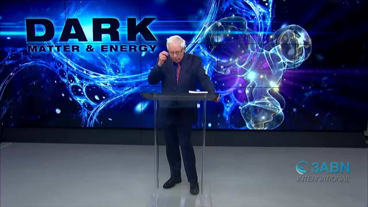 A man in a suit stands at a clear podium, adjusting his glasses. Behind him, a screen displays the words "DARK MATTER & ENERGY" against a swirling, blue background, with the 3ABN International logo in the corner.
