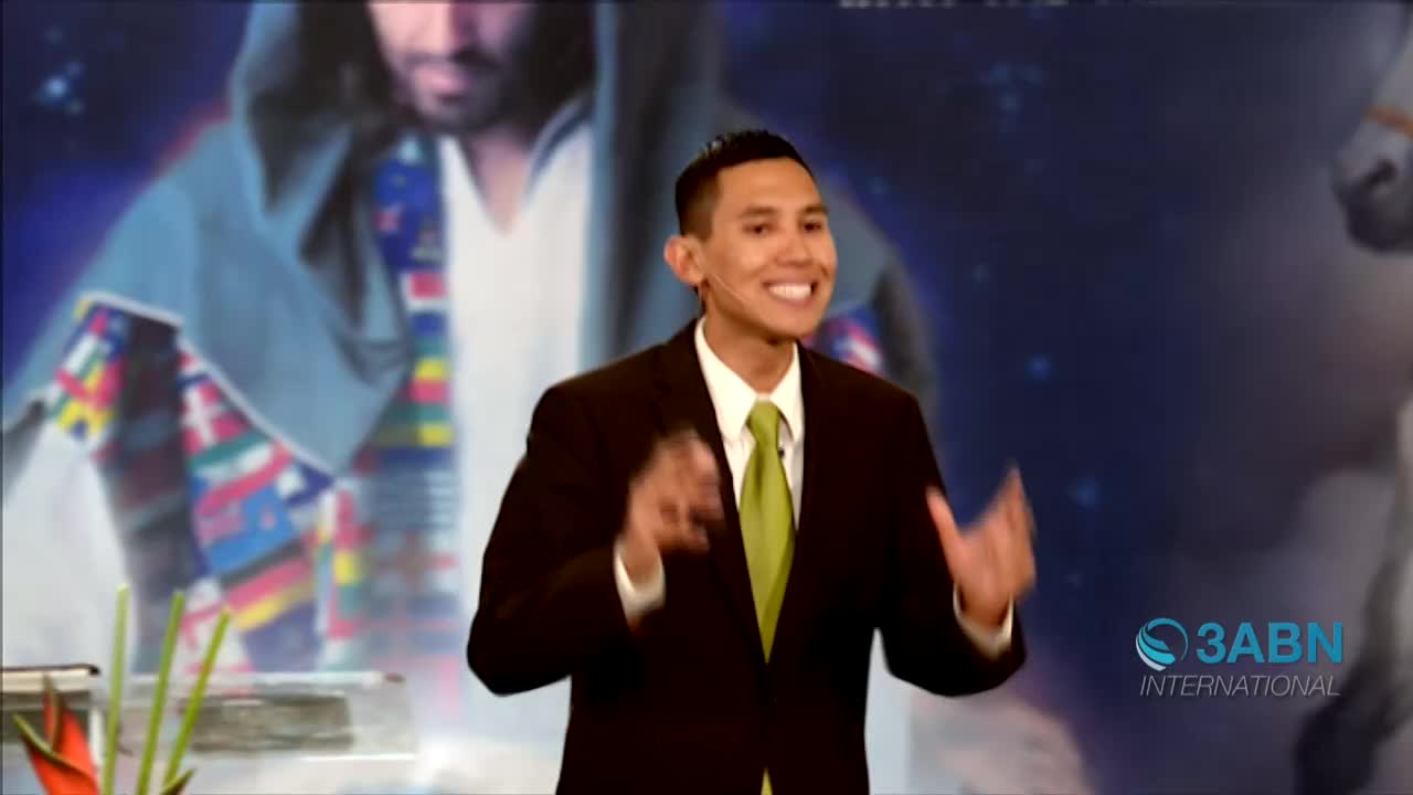 A man in a suit, with a green tie, is speaking animatedly, gesturing with his hands. Behind him, a backdrop depicts a religious figure and a starry night, with the 3ABN International logo visible.
