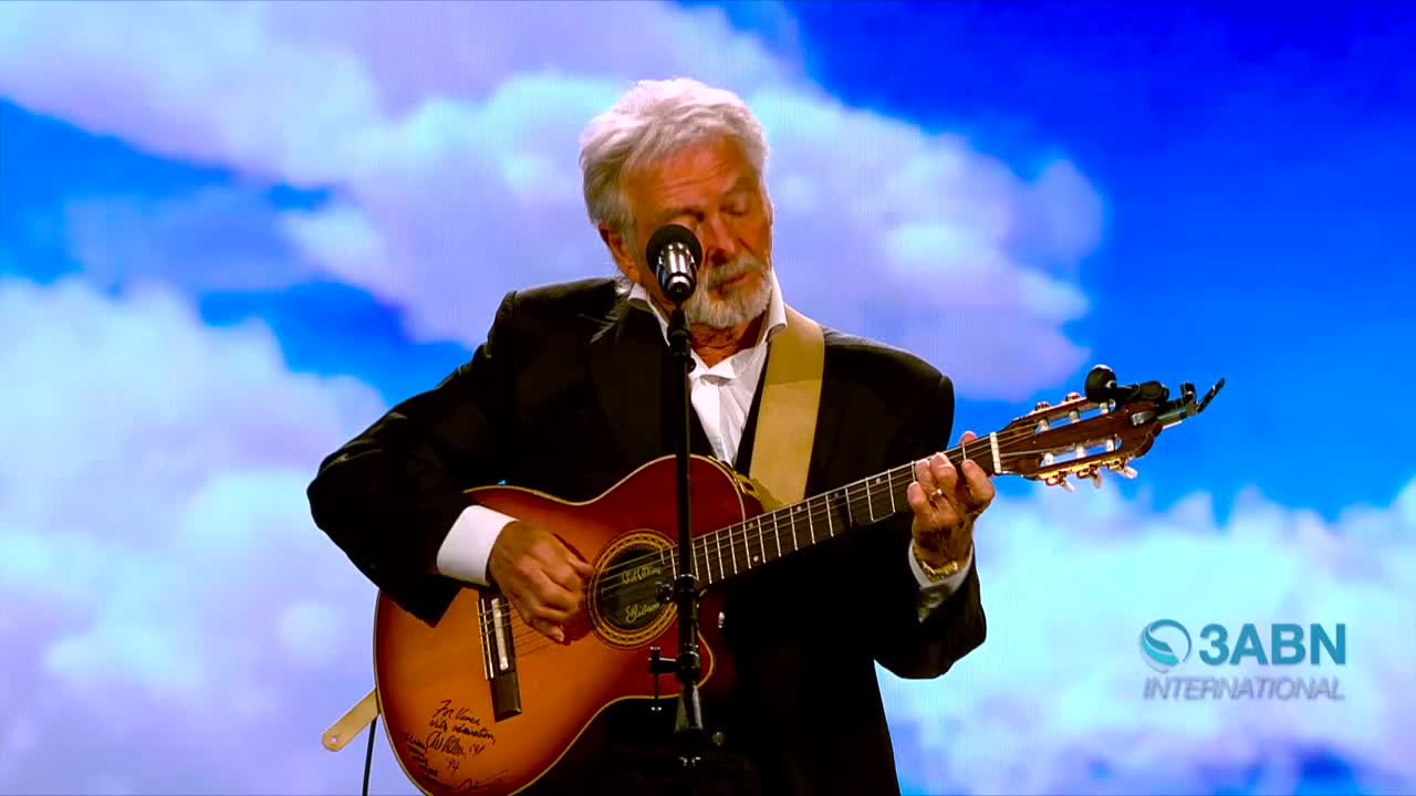 A man in a suit strums a guitar, his fingers dancing over the frets. He's singing into a microphone, the 3ABN International logo visible in the corner.
