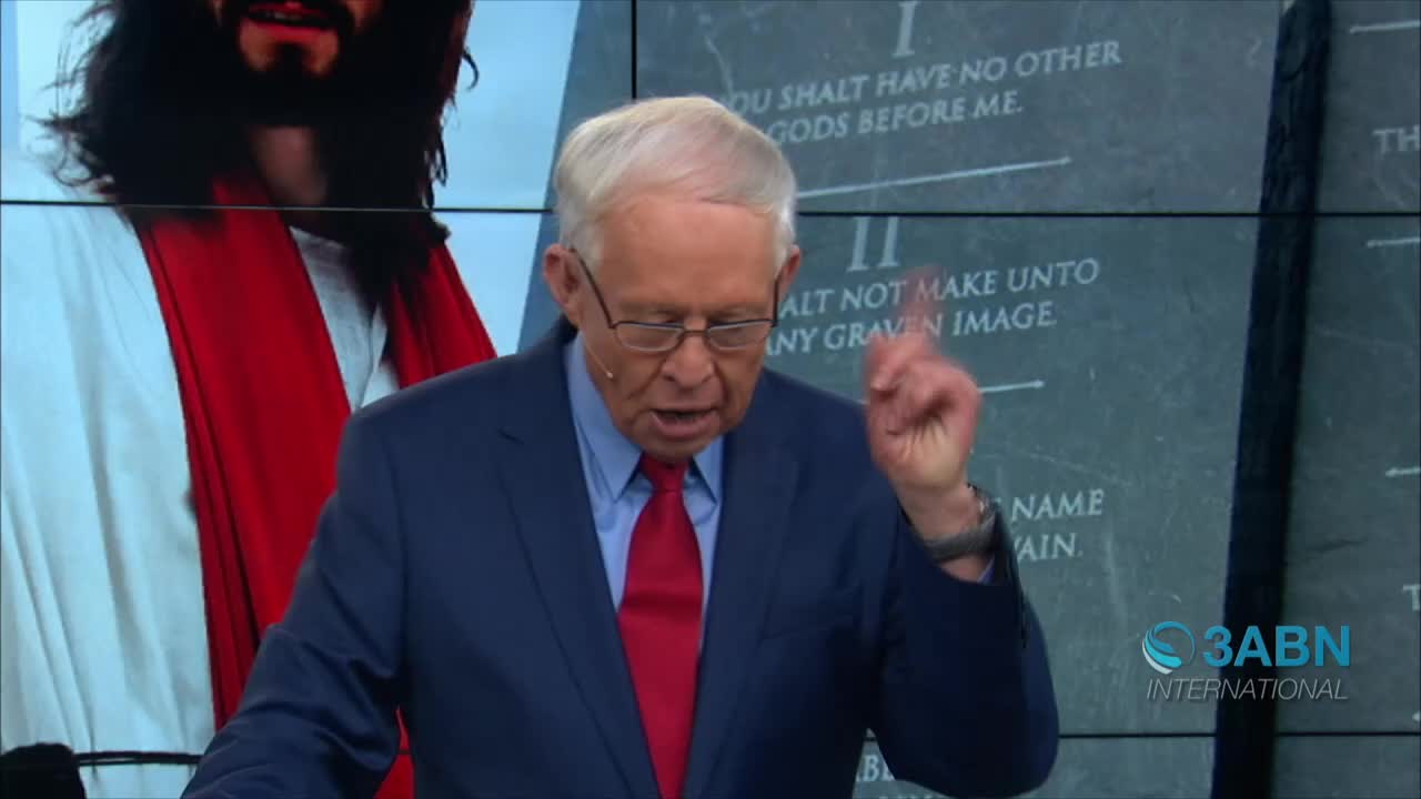 A man in a blue suit and red tie gestures emphatically, pointing towards a screen displaying the Ten Commandments. Behind him, a large image of Jesus in a white robe and red sash is visible.
