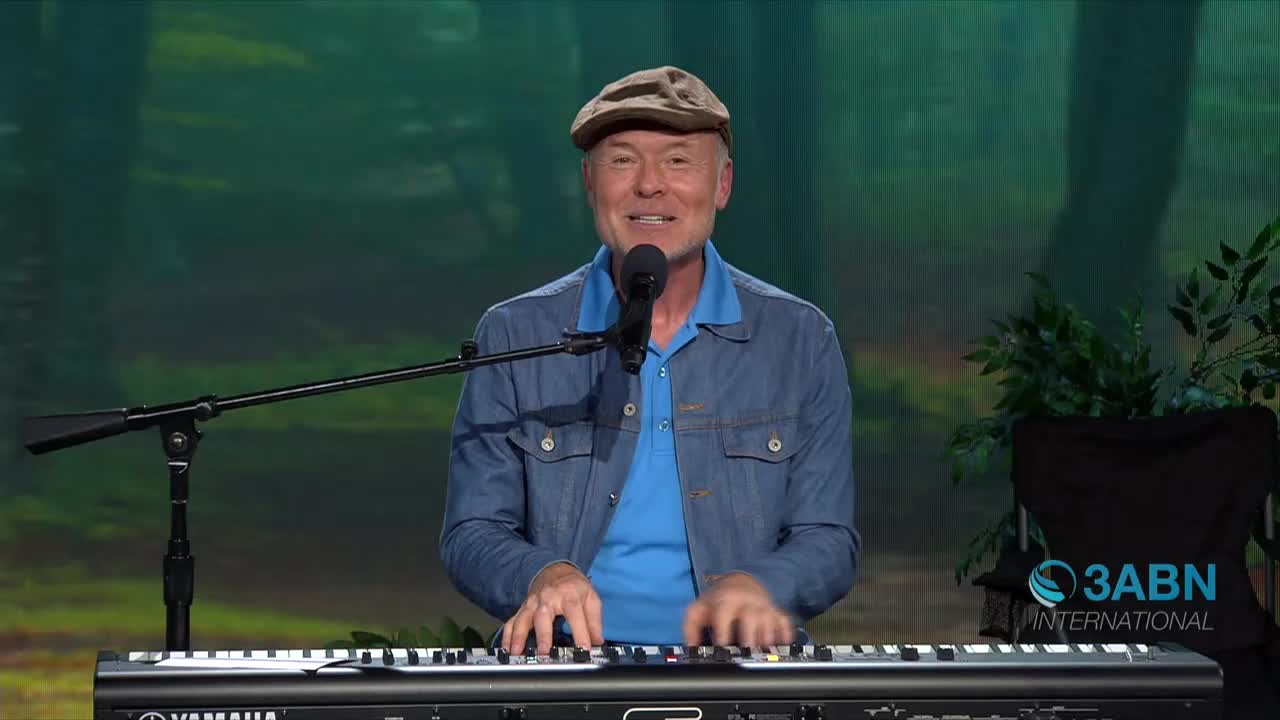 A man in a cap and denim jacket plays a keyboard, his fingers moving across the keys. He smiles at the camera, positioned in front of a backdrop for 3ABN International.
