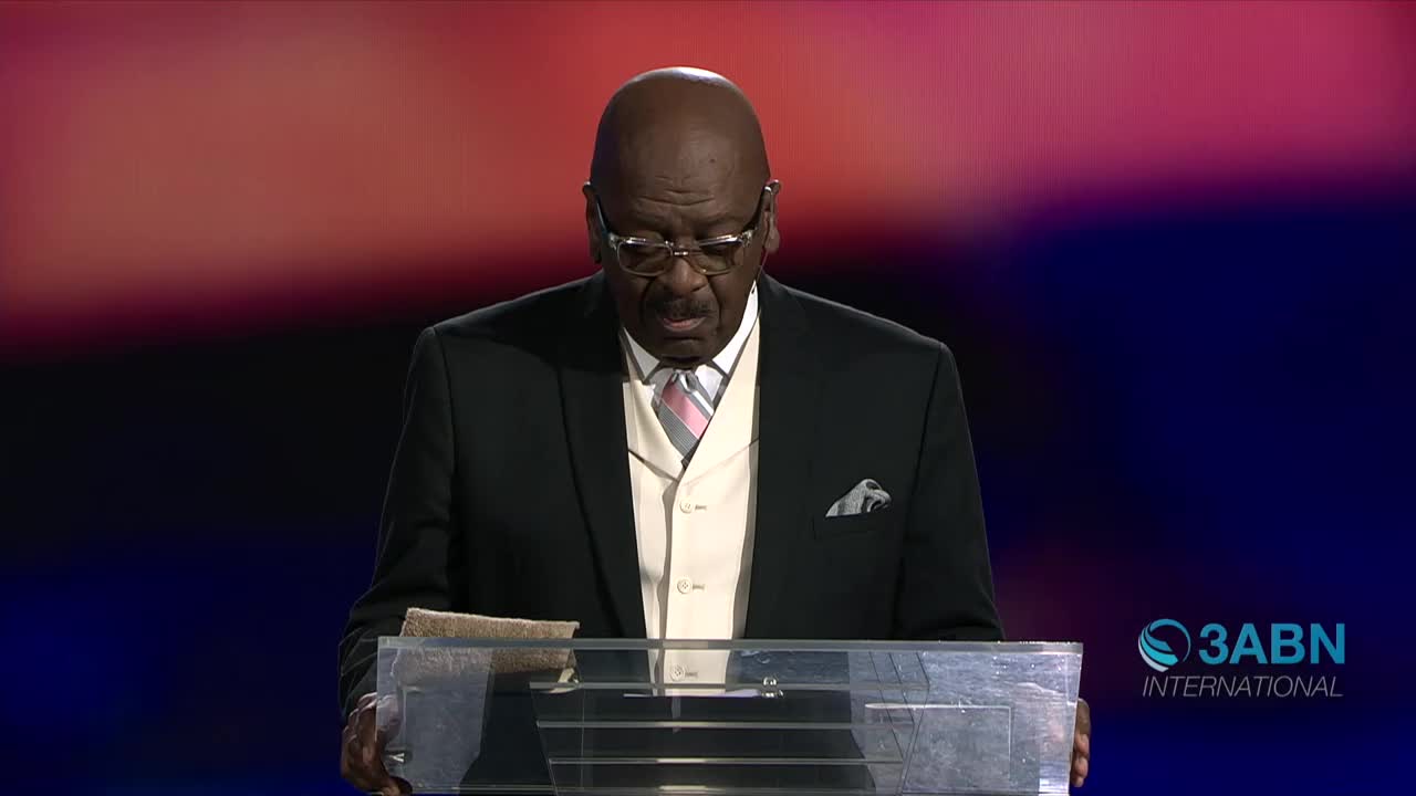 A man in a dark suit and glasses stands at a podium, reading from a book. The 3ABN International logo is visible in the corner.

