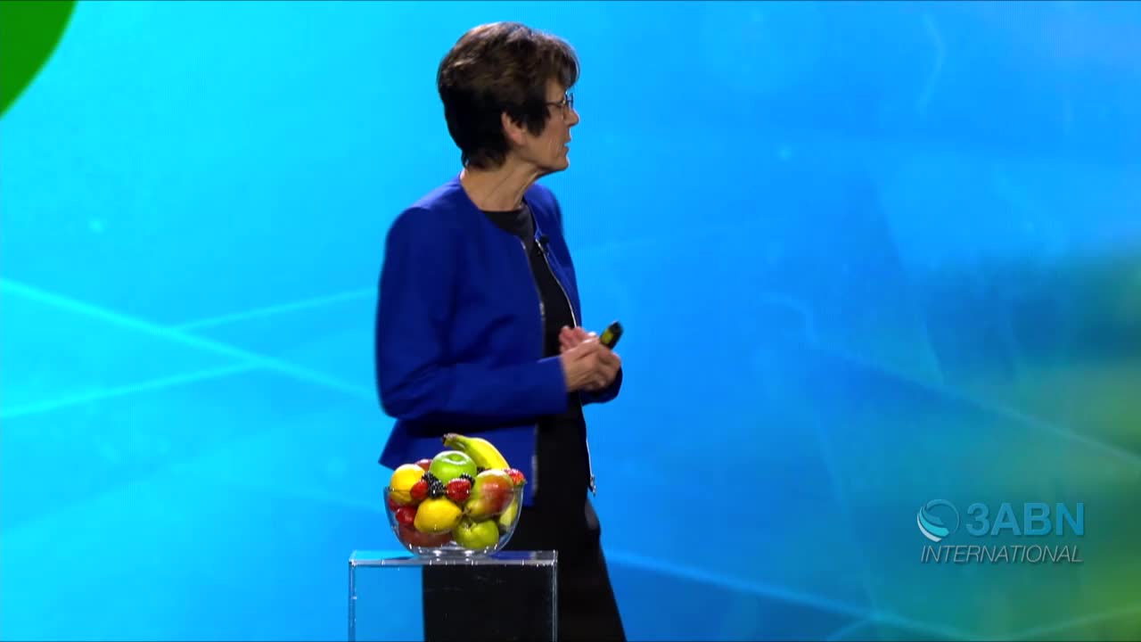 A woman in a blue jacket stands beside a glass bowl filled with fruit, likely on the set of 3ABN International. She holds a small microphone and appears to be addressing an audience.
