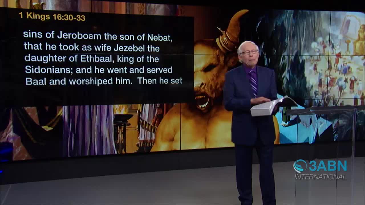 A man in a suit stands before a screen, holding a book open in his hands. Behind him on the screen, text from 1 Kings is displayed alongside an illustration of a horned figure.

