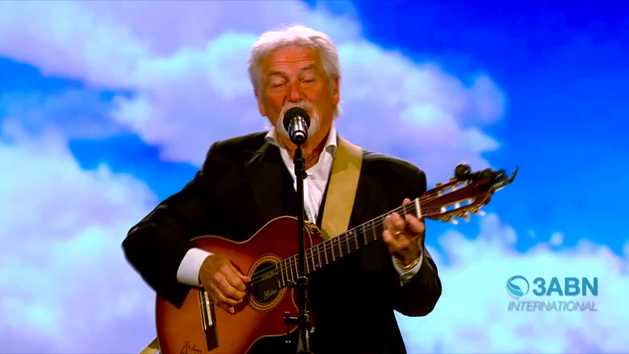 A man in a suit strums a guitar, his voice echoing from the microphone. Behind him, a sky of blue and white clouds fills the backdrop, and the 3ABN International logo is visible.
