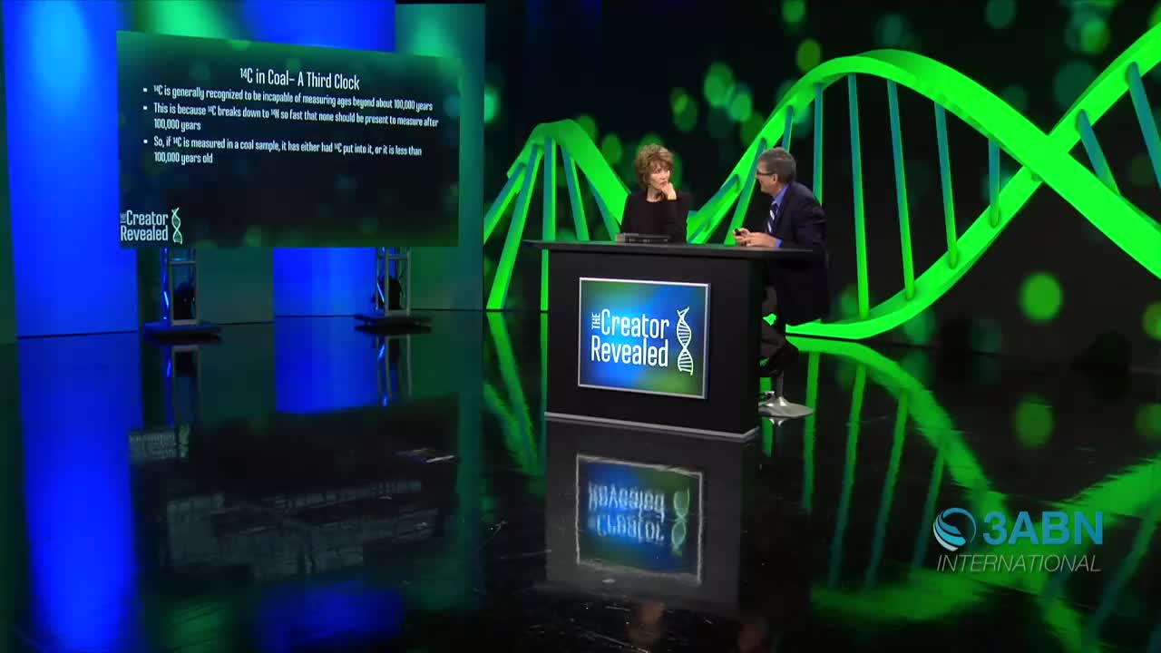 Two people sit at a desk, engaged in a discussion on a set for *The Creator Revealed* on 3ABN International. Behind them, a large screen displays text about carbon dating, with a DNA-like structure as a backdrop.
