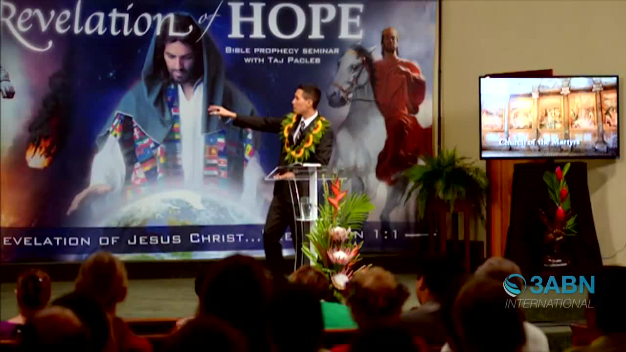 A man in a suit and lei gestures towards a screen, speaking from a podium in front of a large backdrop. The 3ABN International logo is visible, and the presentation is about the Revelation of Jesus Christ.
