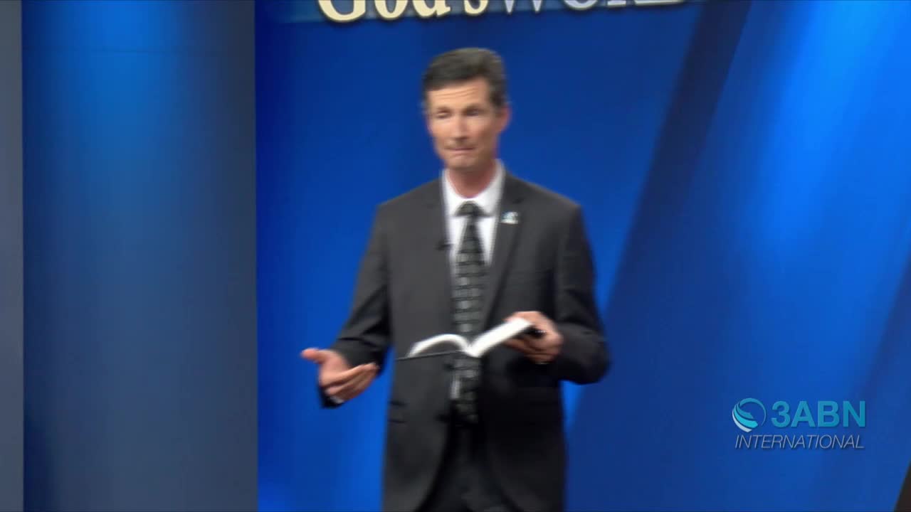 A man in a suit stands center stage, holding a small book open in his hands. He gestures with his other hand as he speaks, likely on a program for 3ABN International.
