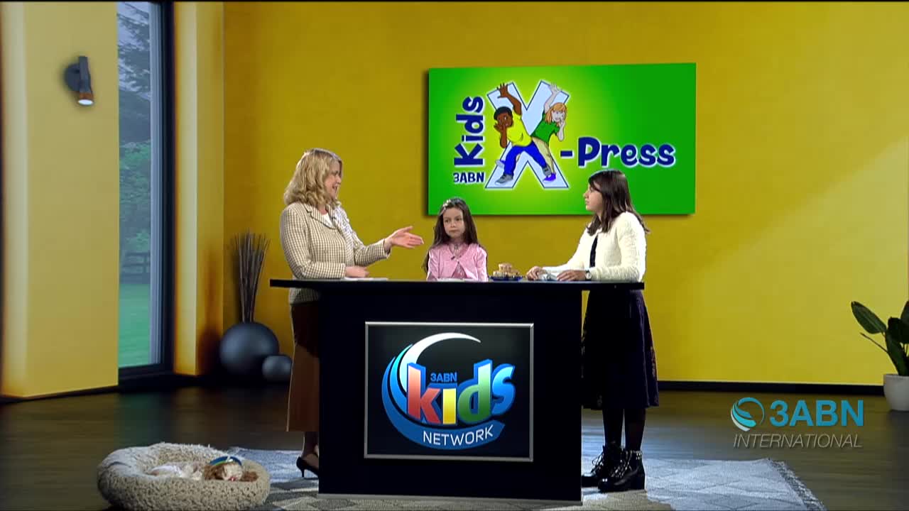 On the set of 3ABN Kids X-Press, a woman in a blazer gestures towards two young girls seated at a desk. A small dog sleeps peacefully in a bed on the floor.
