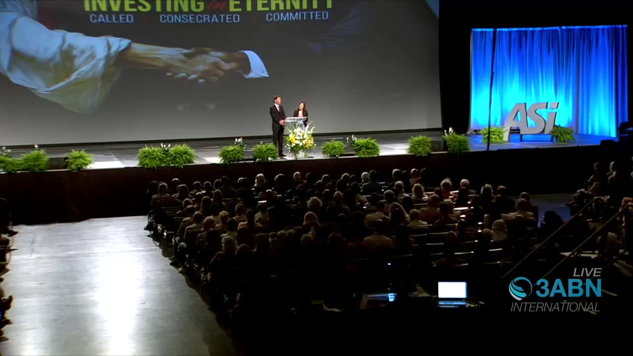 On a stage, two people stand behind a podium, addressing a large audience. A giant screen displays the words "Investing in Eternity" as 3ABN International broadcasts live.
