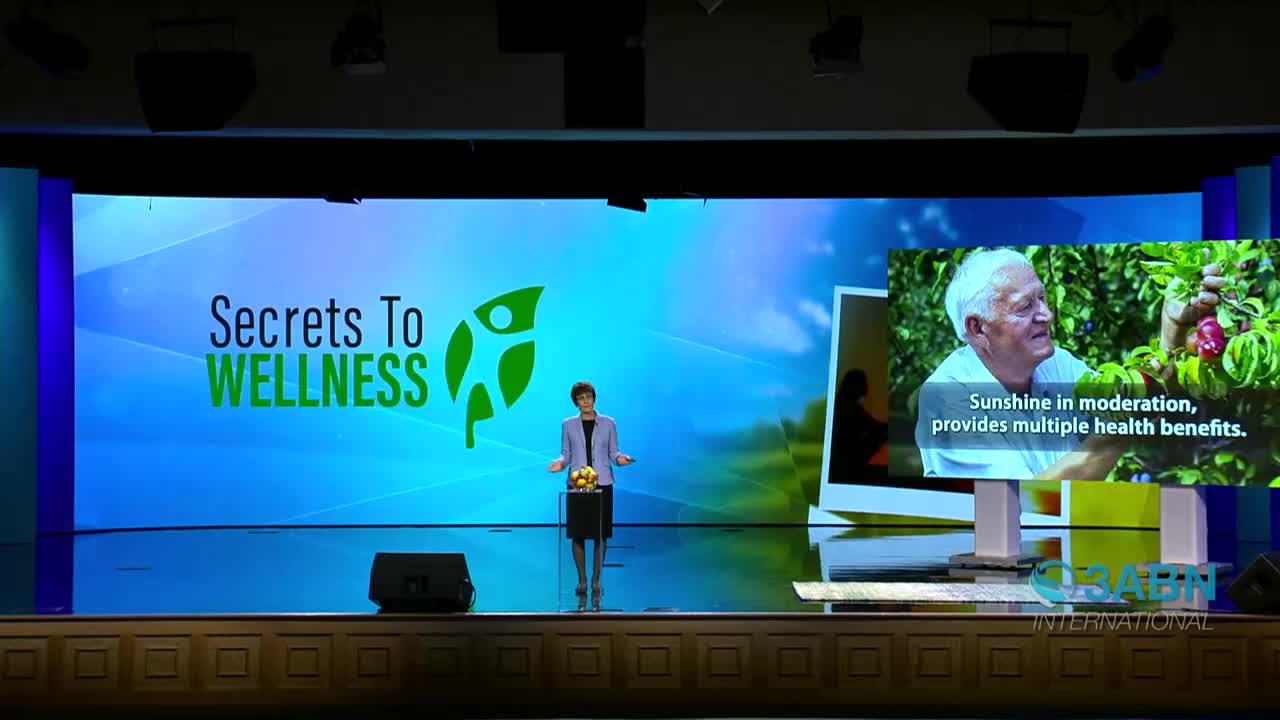 A woman in a blazer stands on a stage, gesturing towards a large screen displaying the words "Secrets to Wellness." Beside her, a smaller screen shows a man picking fruit, with text about the benefits of sunshine.
