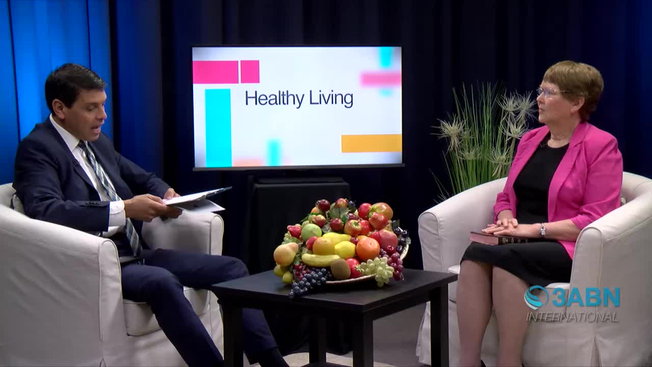 A man in a suit, holding papers, speaks to a woman in a pink blazer. A "Healthy Living" graphic is visible on the screen behind them, and the 3ABN International logo is on the bottom right.
