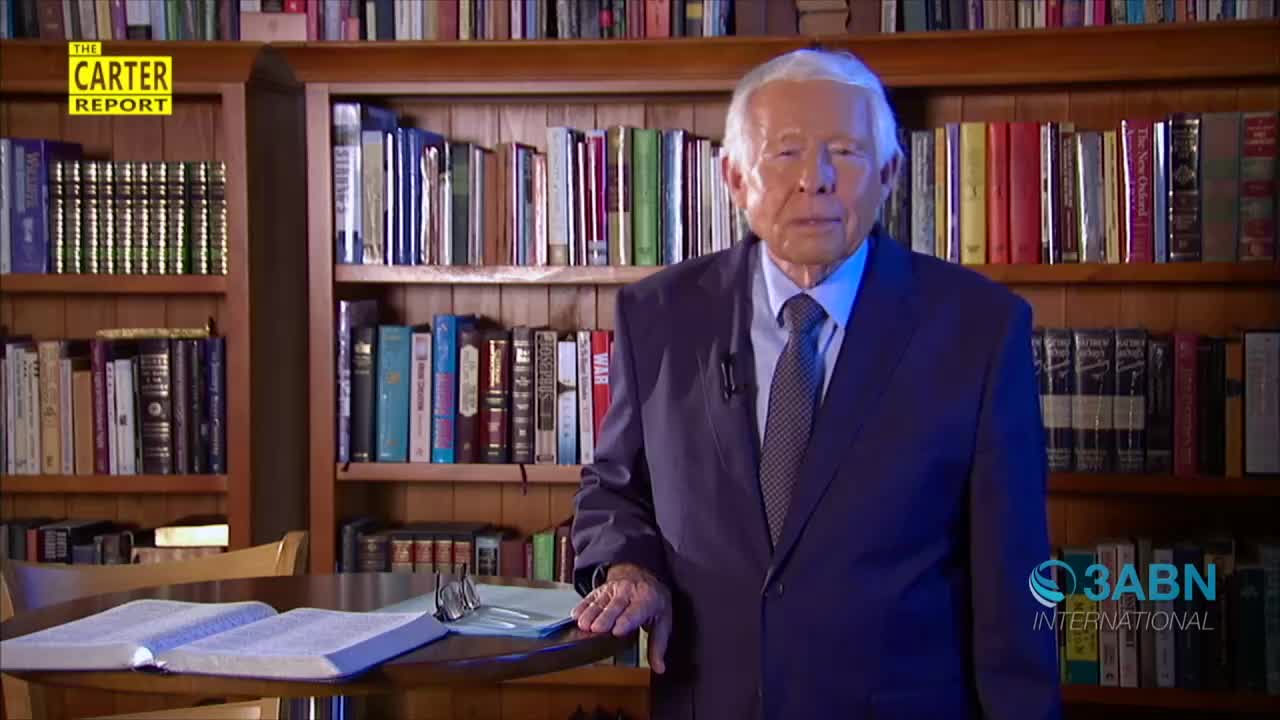 A man in a navy suit stands in front of a bookshelf, addressing the camera on a 3ABN International broadcast. His hands rest on a table with an open book and a pair of glasses.
