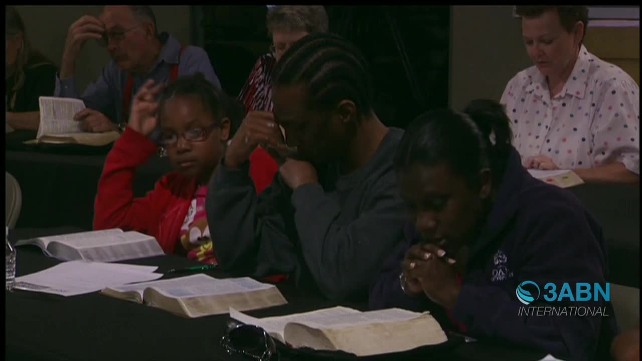 People sit at a table, heads bowed, hands clasped in prayer, with open Bibles in front of them. The 3ABN International logo is visible in the corner.
