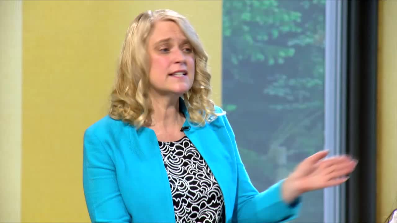A woman in a bright blue jacket gestures with her right hand, speaking animatedly. Her hair is curled, and she's wearing a black and white patterned top.
