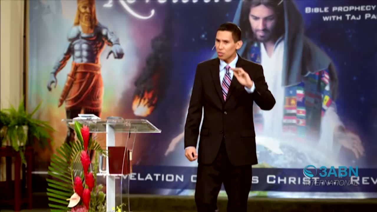 A man in a suit gestures with his right hand, speaking from behind a clear podium. Behind him, a large backdrop from 3ABN International depicts a biblical scene.
