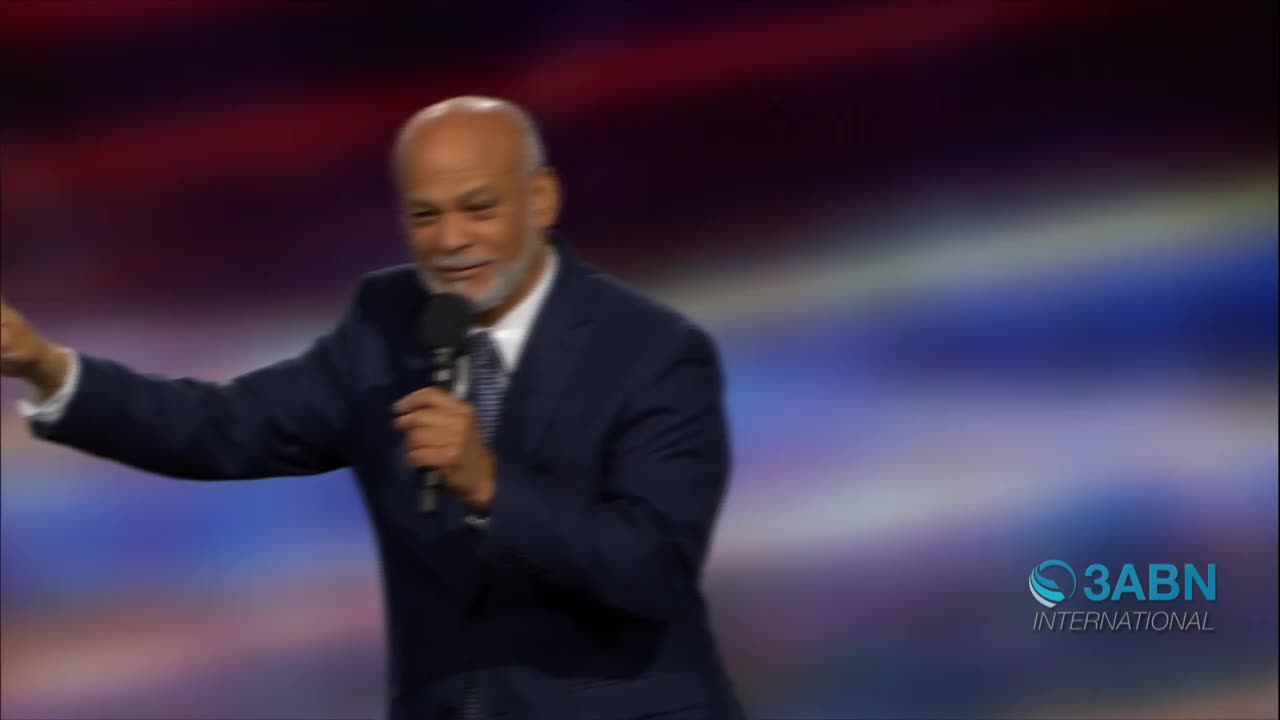 A man in a dark suit gestures with his left arm, holding a microphone in his right hand. The 3ABN International logo is visible in the lower right corner.
