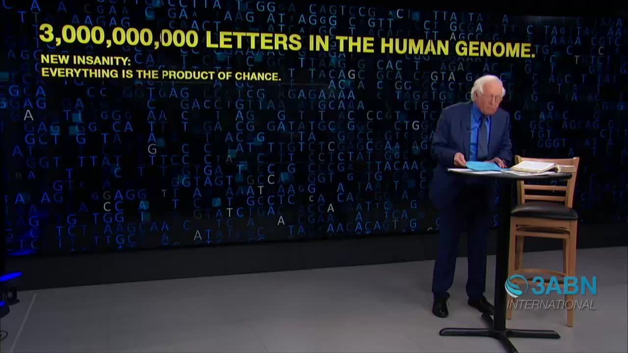 A man in a blue suit stands beside a small table, holding papers as he looks down at them. Behind him, a screen displays text about the human genome, with the 3ABN International logo visible in the corner.
