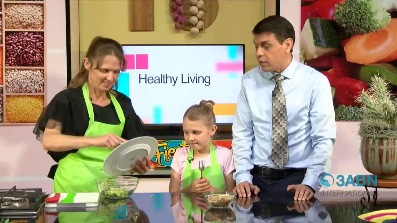 A woman in a green apron holds a plate over a bowl of green food as a young girl with a fork looks on. The man in a blue shirt and tie watches them on the set of 3ABN International's "Healthy Living" program.

