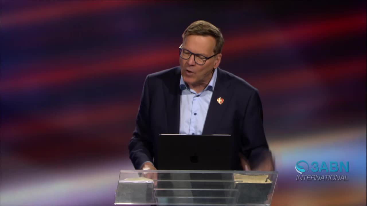 A man in a suit and glasses speaks into a microphone, standing behind a podium with a laptop. The 3ABN International logo is visible in the corner.
