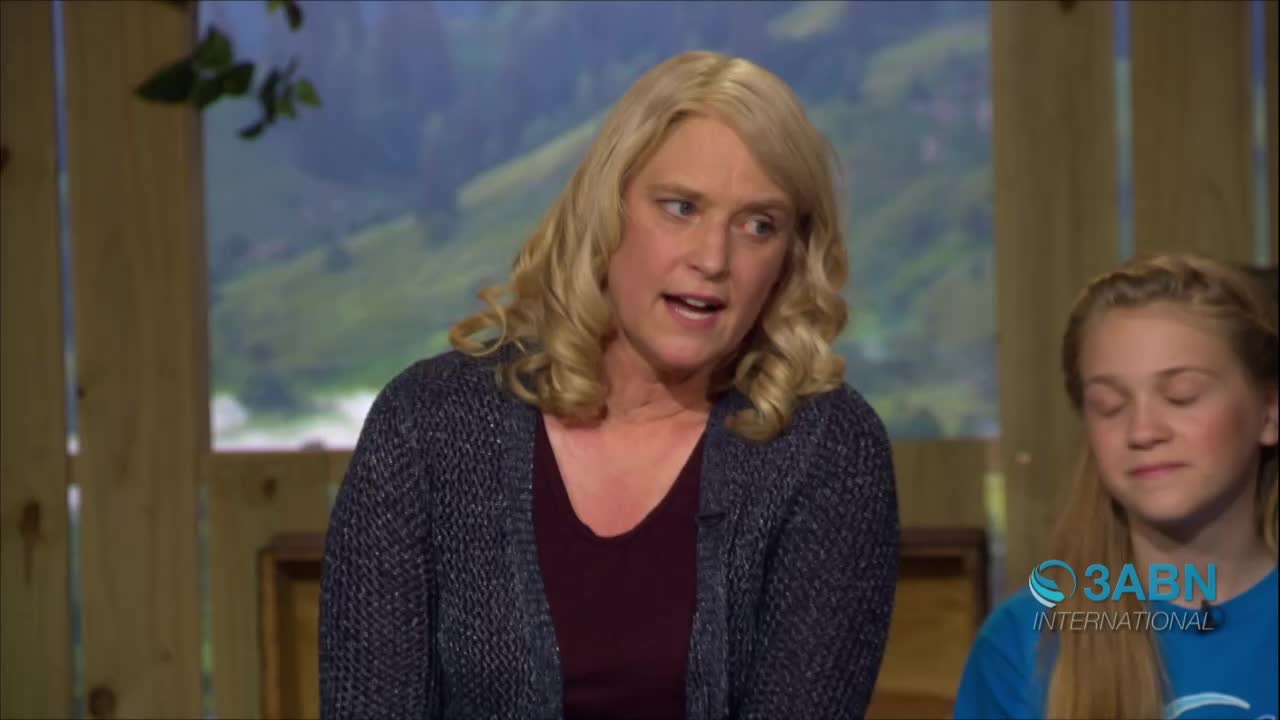A woman with blonde, wavy hair speaks animatedly, wearing a dark cardigan over a burgundy top. Beside her, a younger girl in a blue shirt bearing the 3ABN International logo listens intently.
