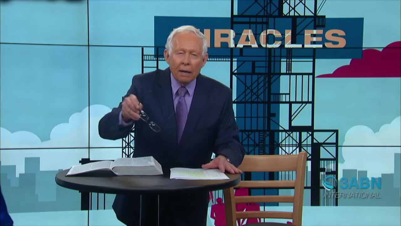 A man in a suit, holding glasses, gestures toward the camera while speaking. Behind him, a large graphic with "MIRACLES" is visible on the set of 3ABN International.
