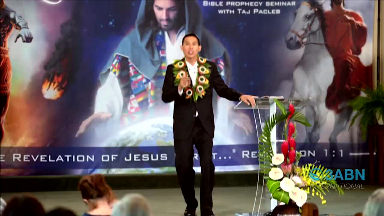 A man in a suit and lei gestures as he speaks from behind a clear podium in front of a large backdrop. The words "Bible Prophecy Seminar" and "3ABN International" are visible, suggesting a religious broadcast from the United States.
