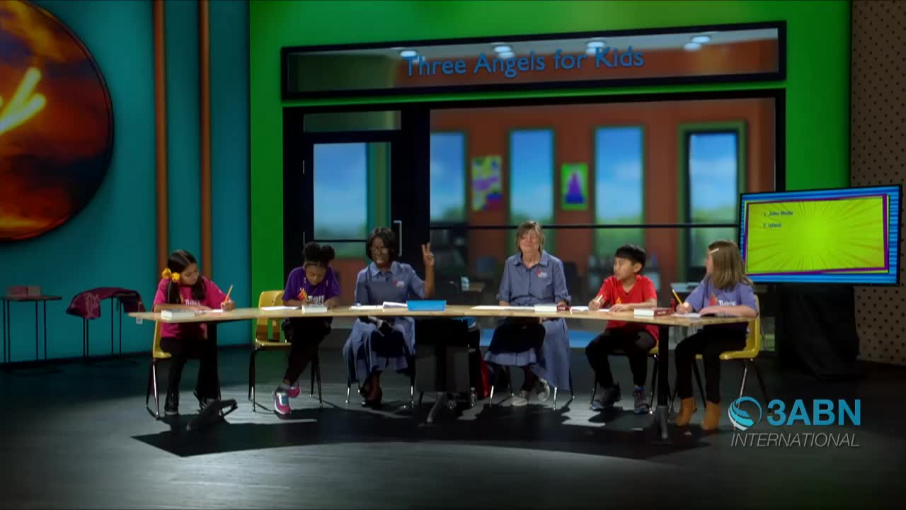 On the set of 3ABN International's "Three Angels for Kids," children are seated at a long table, writing. The woman in the middle is holding up two fingers, and a screen displays a list.
