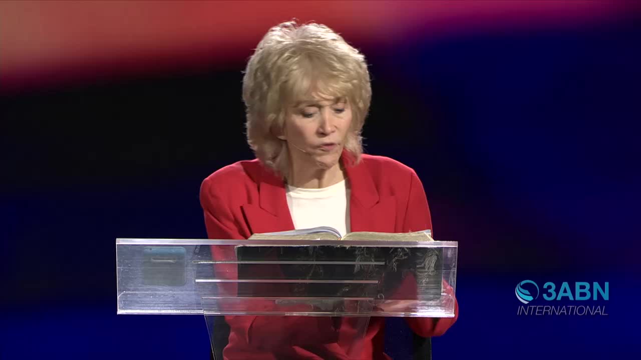 A woman in a red blazer reads from an open book, her eyes moving across the pages. Behind her, the 3ABN International logo is visible against a dark blue backdrop.
