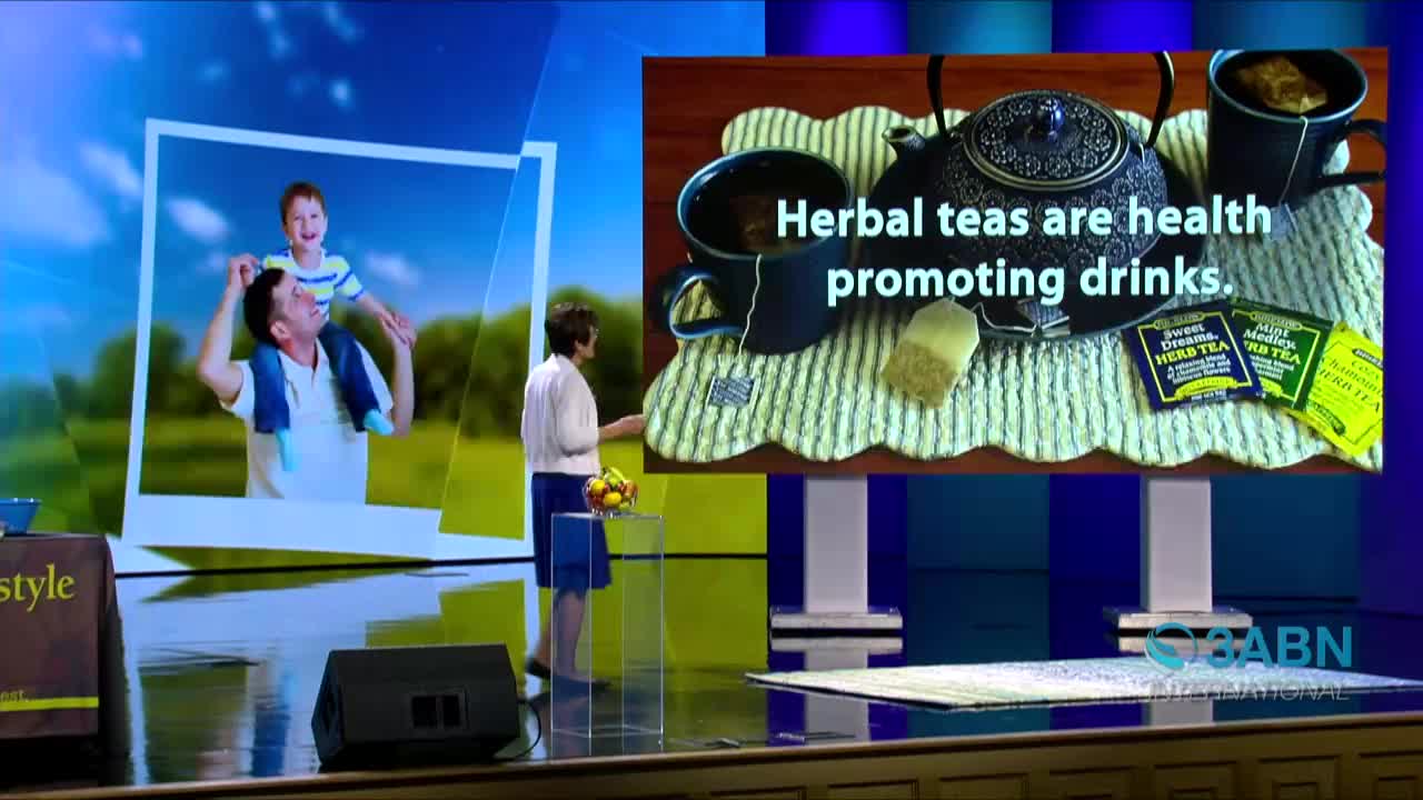 A woman in a white shirt and blue skirt gestures toward a screen displaying images of a father carrying his son and a tea set. The 3ABN International logo is visible at the bottom right.
