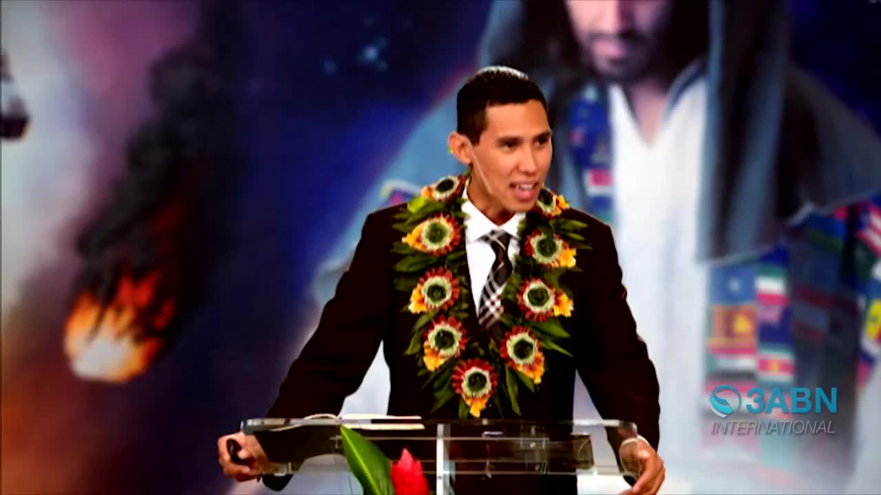 A man in a suit and lei speaks from behind a podium, the 3ABN International logo visible. Behind him, a backdrop of flames and an image of a robed figure adds to the scene's dramatic feel.
