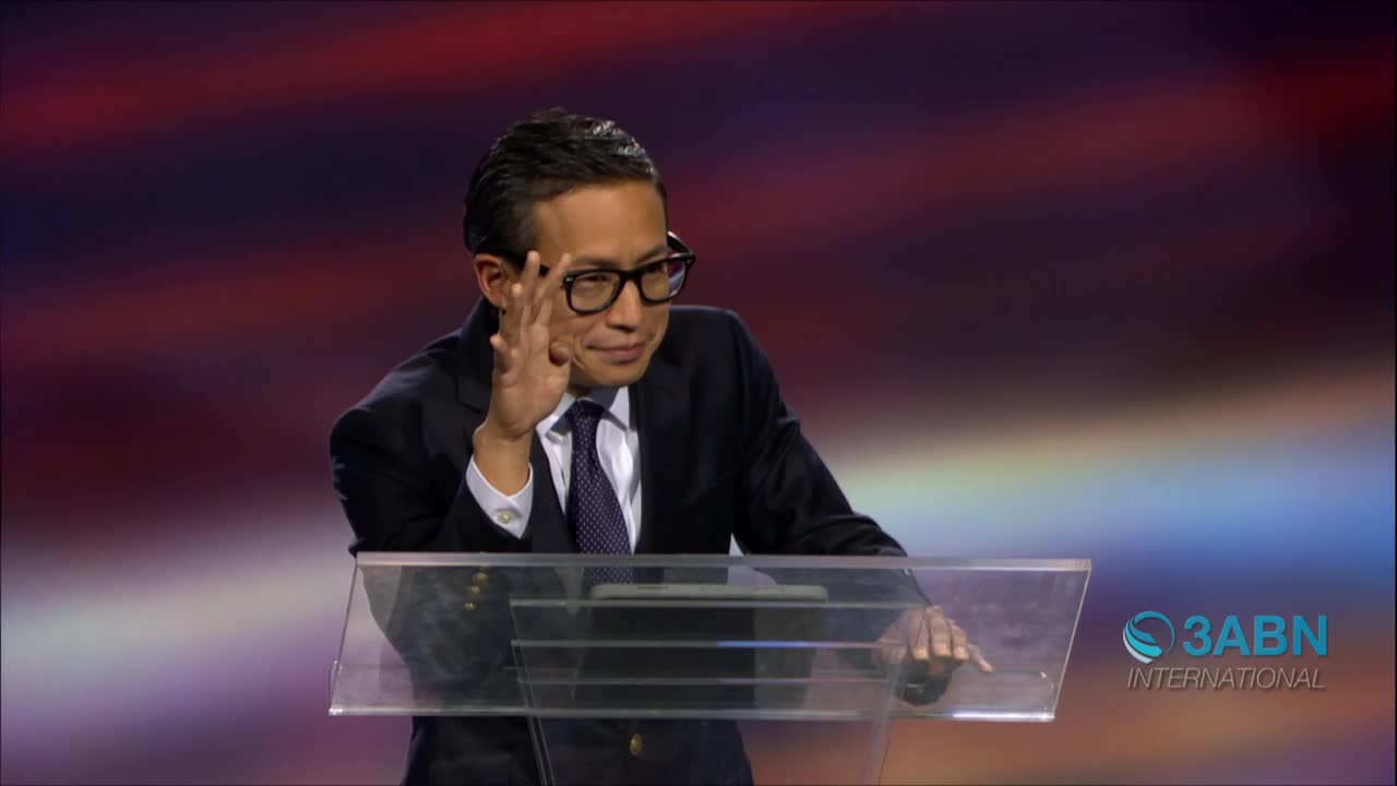 The man in the suit, speaking on 3ABN International, adjusts his glasses with his right hand. He's leaning slightly on the podium, gesturing with his left hand.
