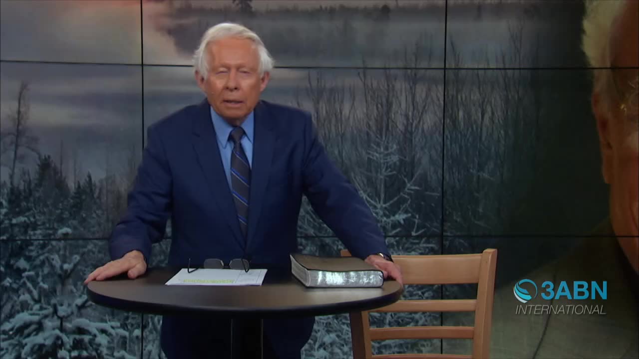 A man in a blue suit stands behind a small table, speaking in front of a backdrop. A Bible rests on the table, next to a pair of glasses and some papers.
