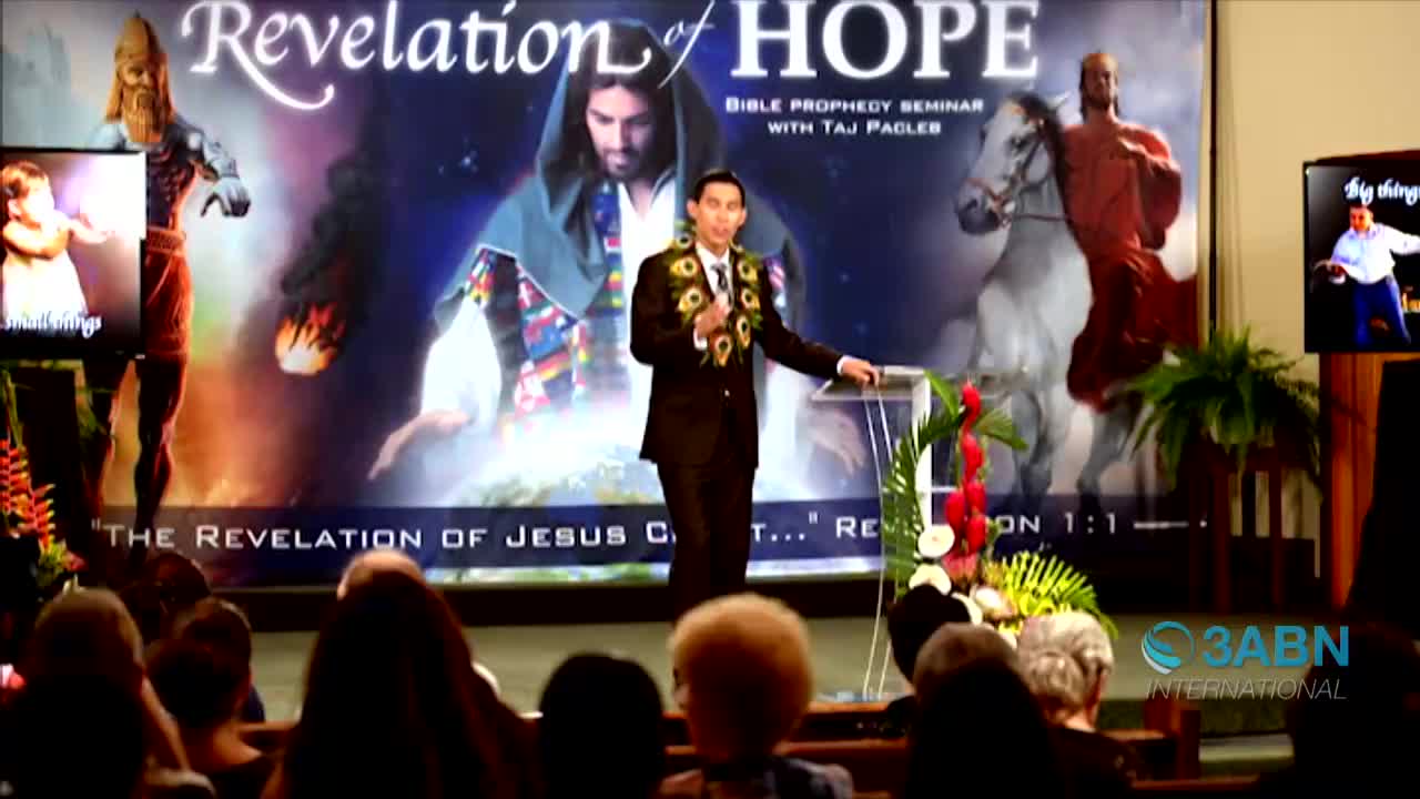 A man in a suit, wearing a lei, stands at a podium and speaks to an audience. Behind him, a large banner advertises a Bible prophecy seminar, and the 3ABN International logo is visible.
