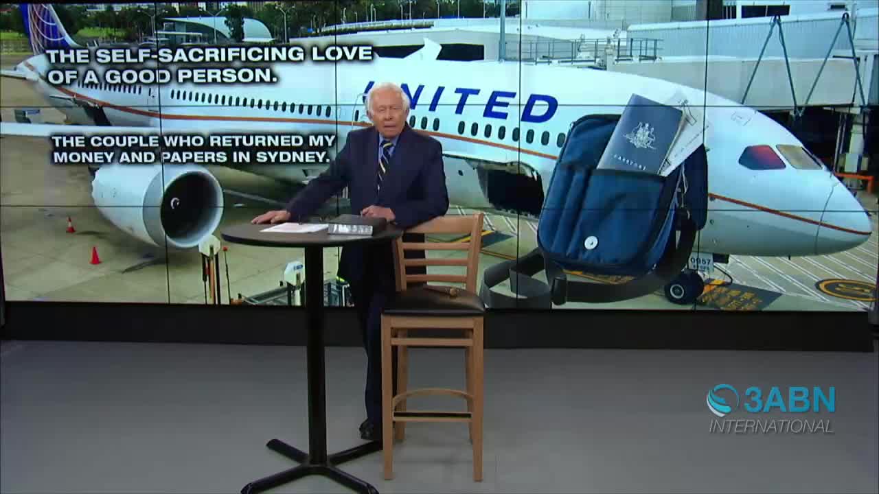 A man in a suit stands beside a small table, gesturing toward a screen displaying a United Airlines plane and a backpack. The 3ABN International logo is visible in the lower right corner.
