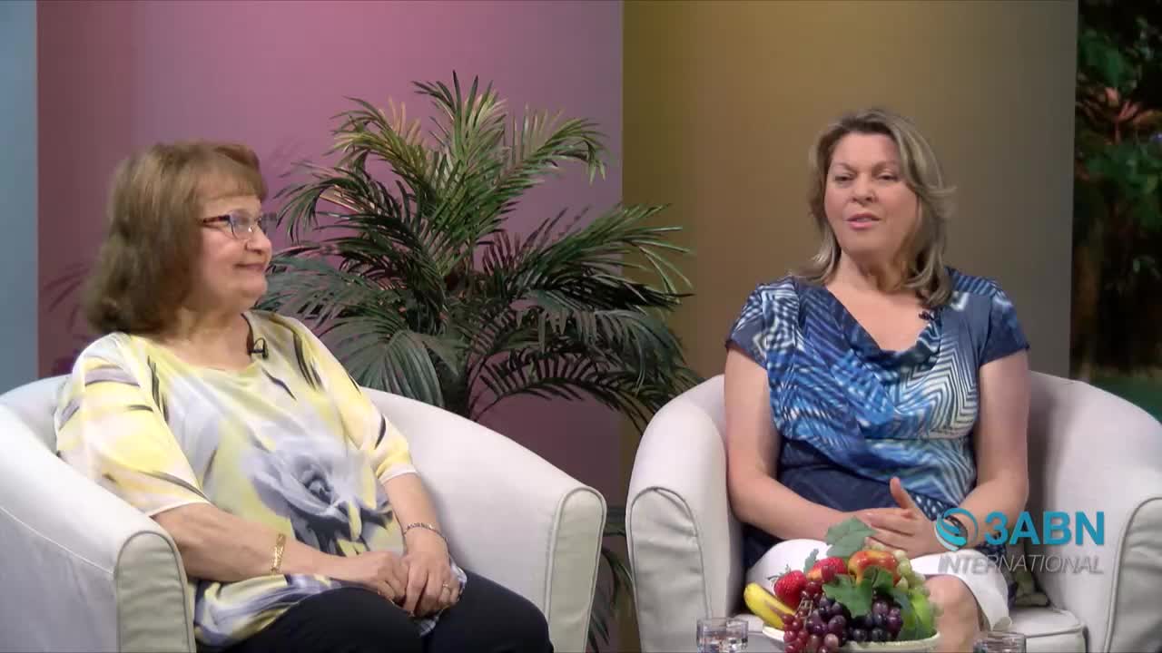 Two women sit in white armchairs, facing forward in a studio setting. The 3ABN International logo is visible on the right, and a bowl of fruit sits on a table between them.
