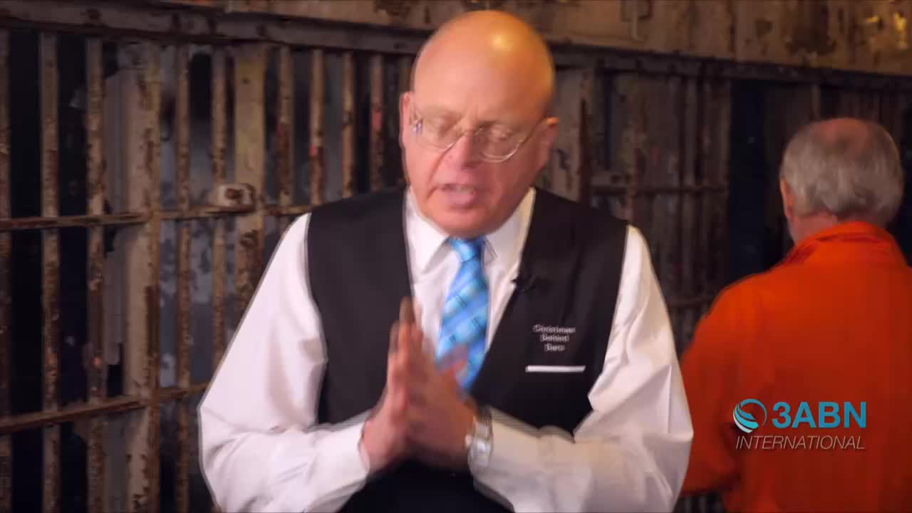 A man in a vest and tie gestures with clasped hands, speaking in front of a rusty jail cell. Another man in an orange jumpsuit stands nearby, and the 3ABN International logo is visible.
