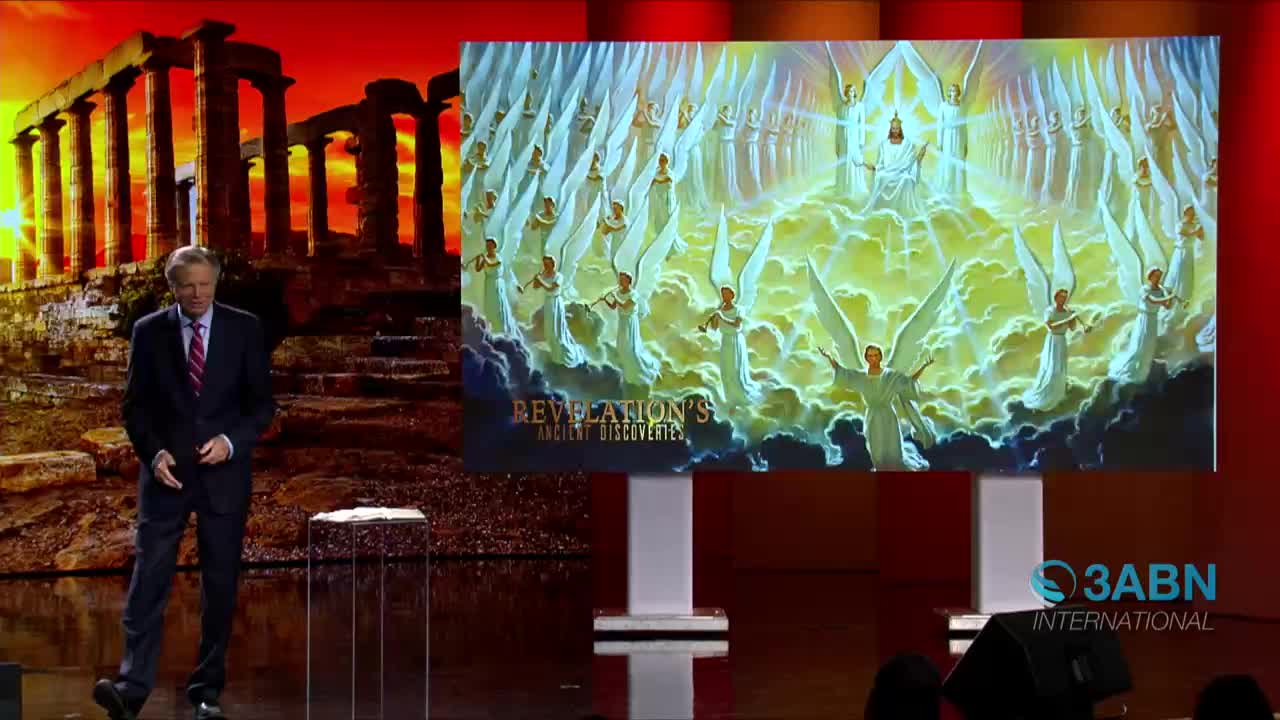 A man in a suit gestures toward a large screen displaying an illustration of angels. The 3ABN International logo is visible on the right, and the backdrop shows a classical building and a sunset.
