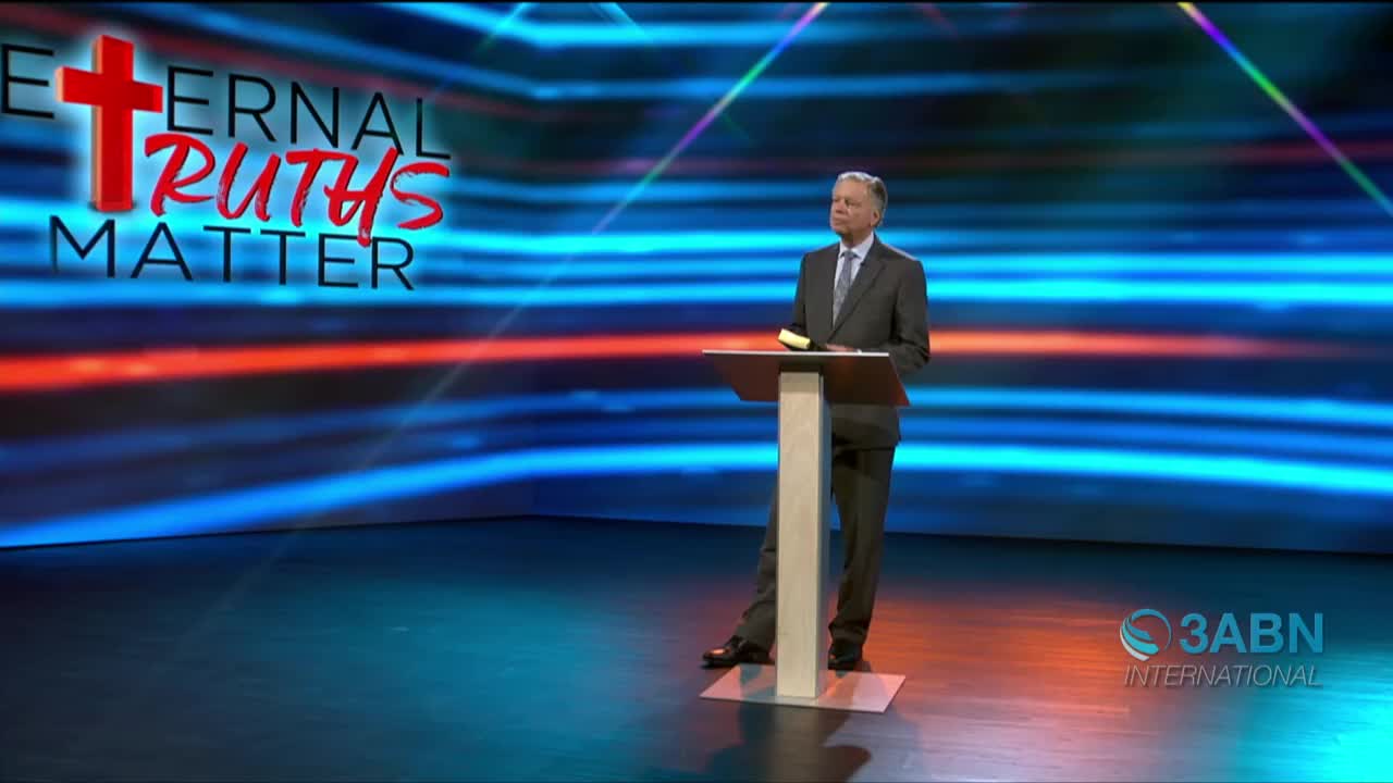 A man in a suit stands behind a podium, a book resting on its surface. Behind him, a backdrop of blue and red streaks fills the screen, with the 3ABN International logo visible in the corner.
