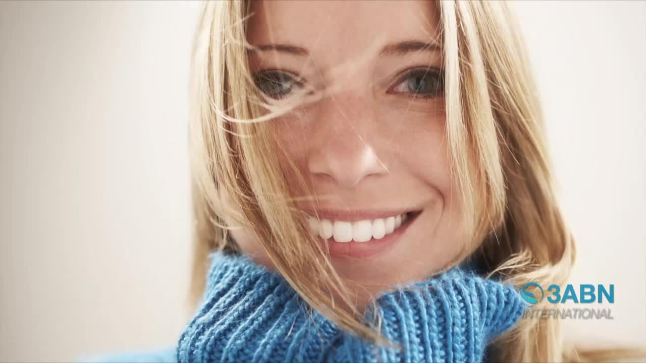 A woman with wind-blown blonde hair smiles widely, revealing a bright, white smile. She's wearing a blue sweater, and the 3ABN International logo is visible in the corner.
