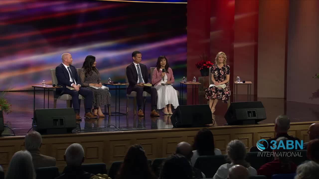 On a stage, six people sit in chairs, with one woman holding a microphone. The 3ABN International logo is visible on the right, and an audience watches from the seats below.
