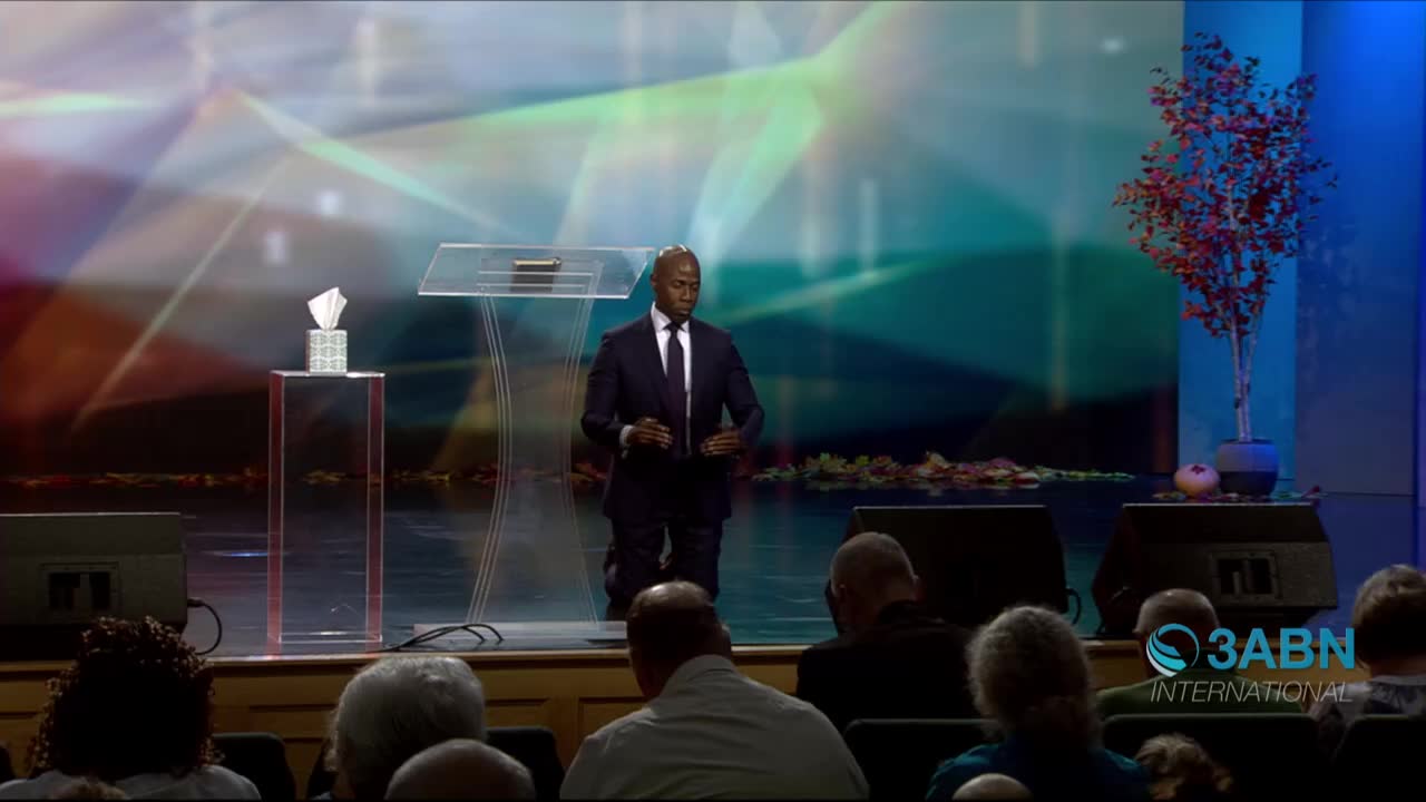 A man in a dark suit kneels on a stage, holding a microphone. Behind him, the 3ABN International logo is visible, and the backdrop is lit with abstract colors.
