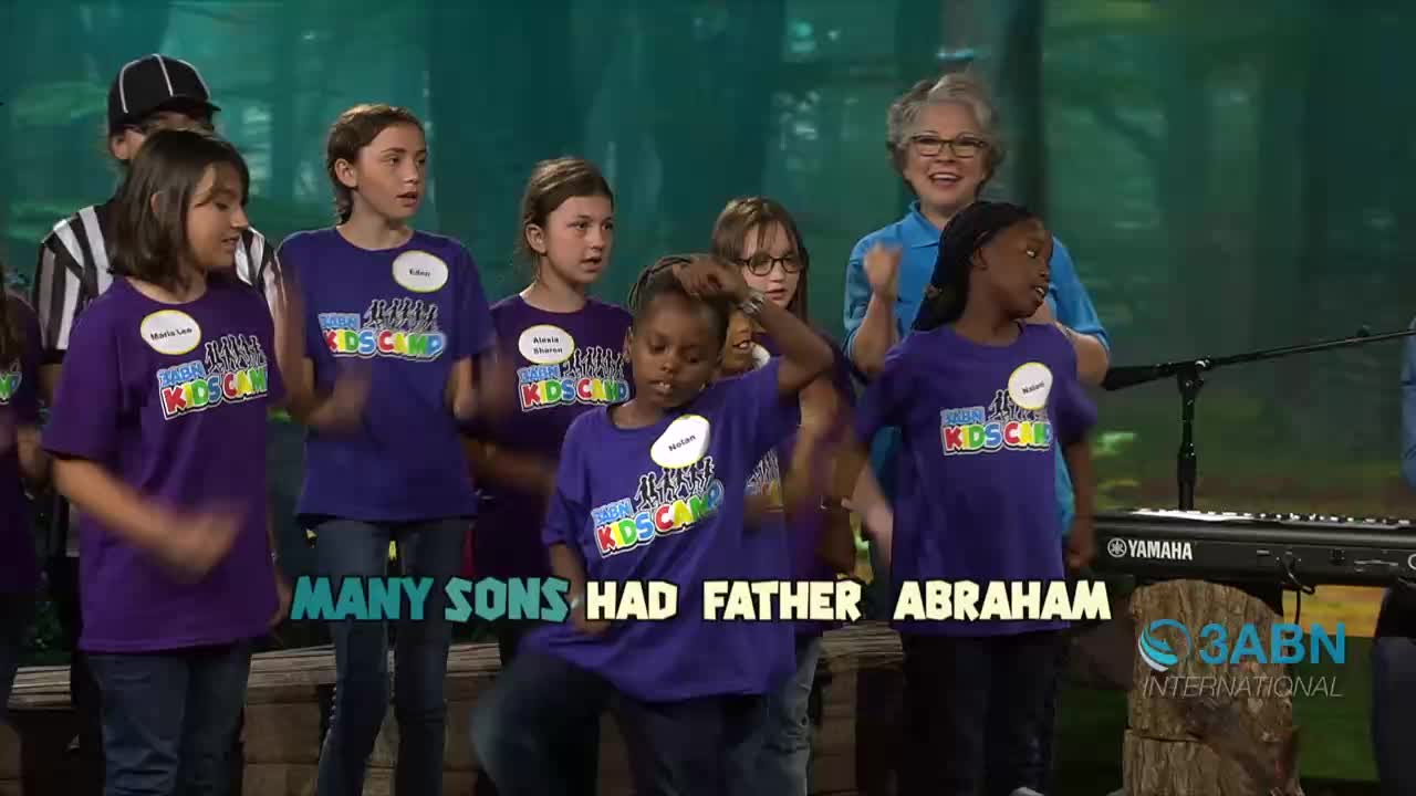 A group of kids in matching purple shirts are singing and moving on a stage, with a woman in glasses and a blue shirt smiling behind them. The words "MANY SONS HAD FATHER ABRAHAM" are displayed in front of them, and the 3ABN International logo is visible.
