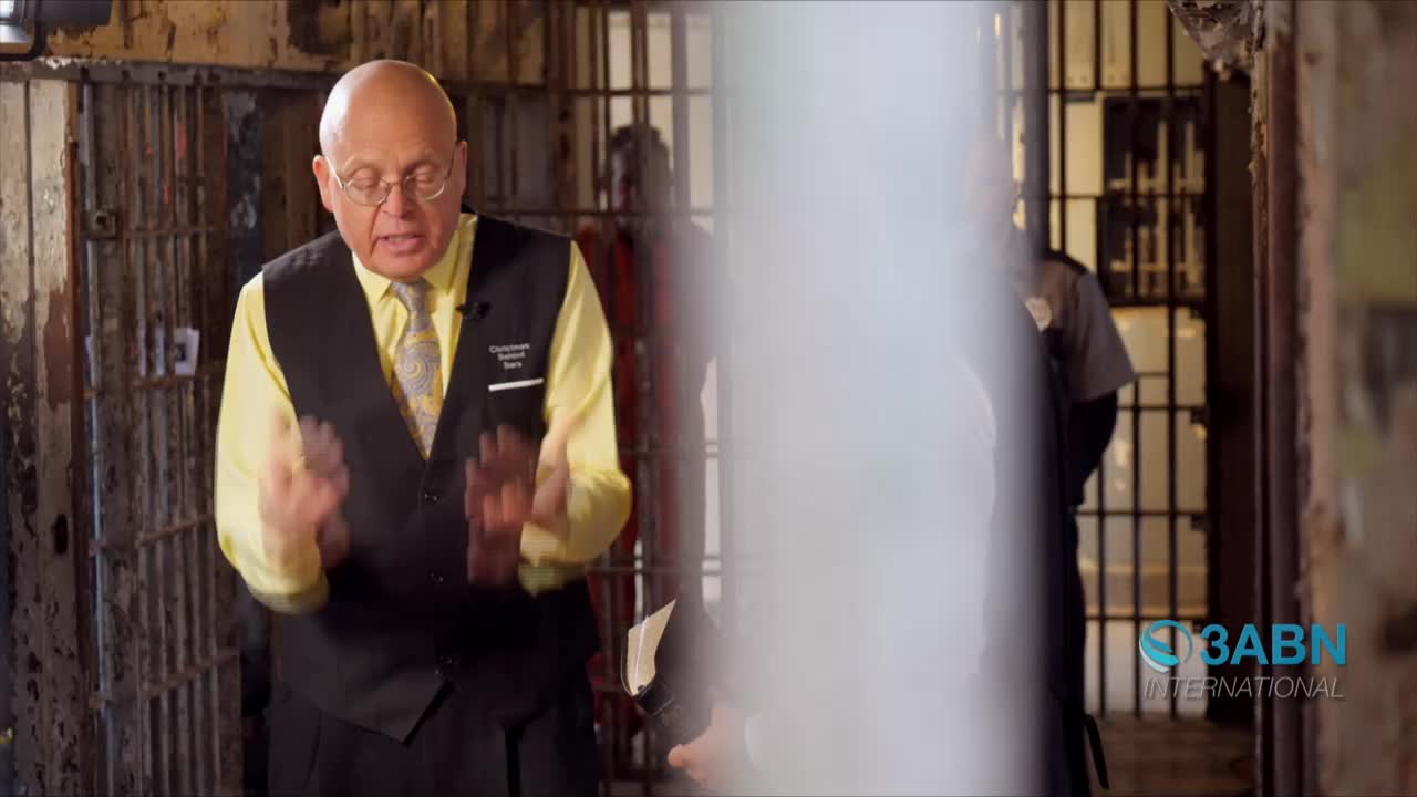 Inside a jail cell, a man in a vest and tie gestures with his hands as he speaks. Behind him, a guard stands in the shadows of another cell.
