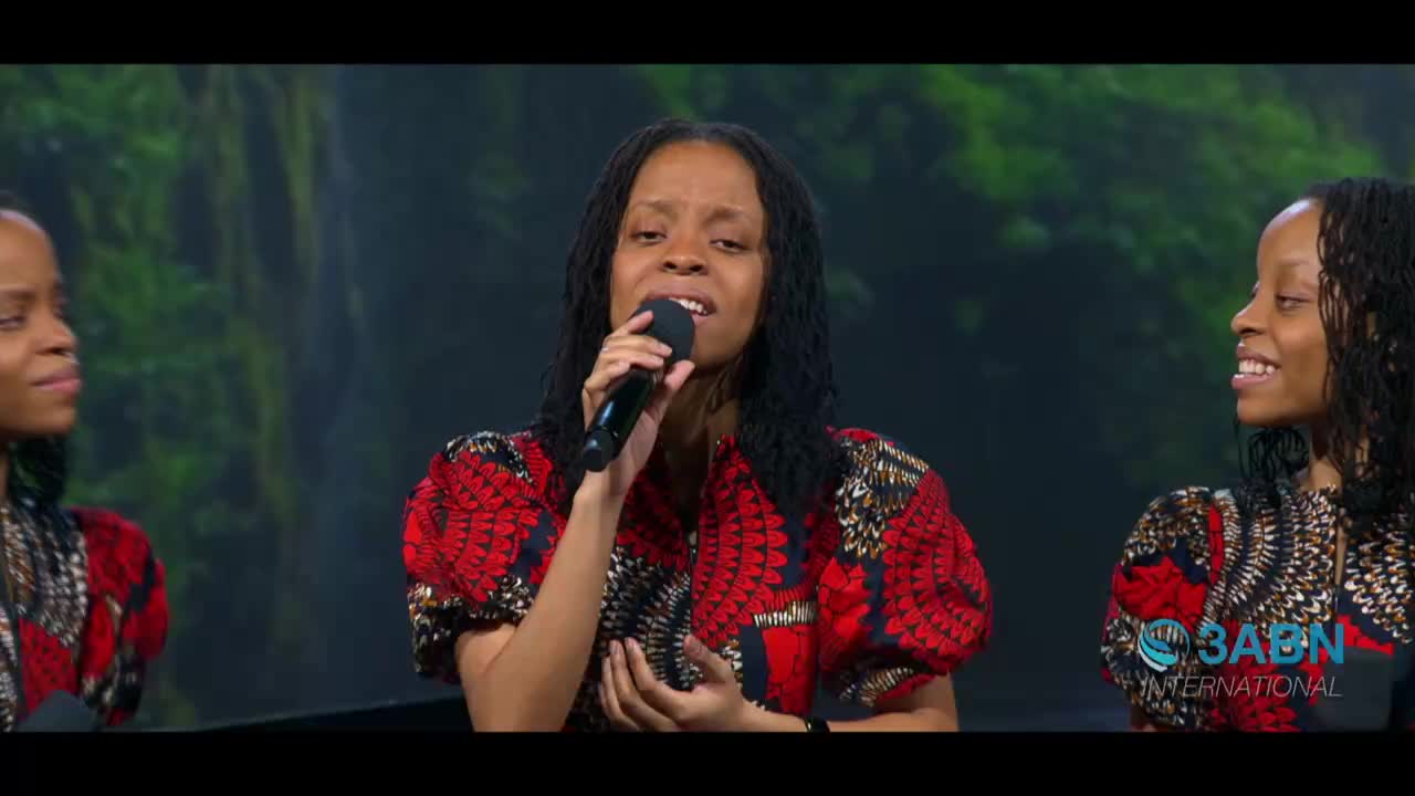A woman in a vibrant red and black patterned top sings into a microphone, her voice carrying across the studio. Flanking her, two other women smile, their matching outfits and hairstyles creating a harmonious visual.
