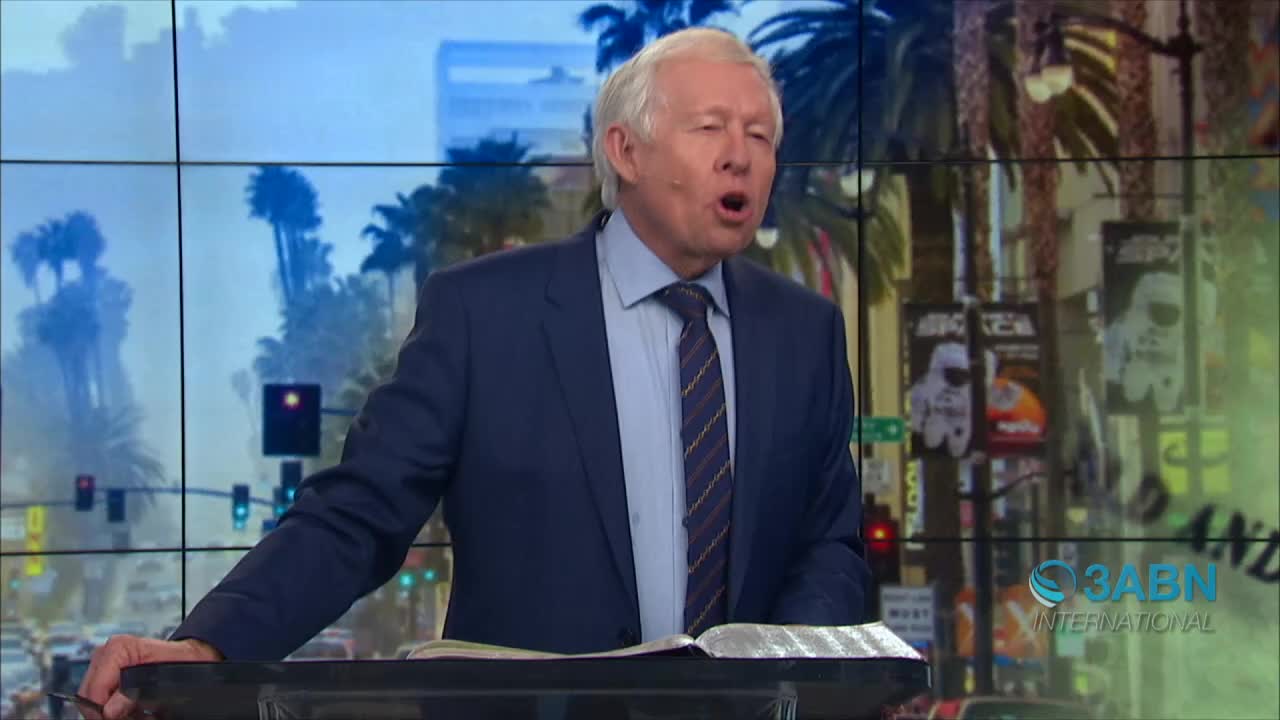 A man in a suit speaks animatedly from behind a lectern, his mouth open. Behind him, a screen displays a street scene, and the 3ABN International logo is visible.
